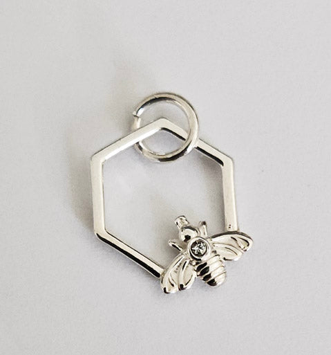 Närbild på ett silversmycke format som en sexkantig hexagon. I botten av ramen sitter ett litet, detaljerat bi i silver med en klar sten på ryggen.