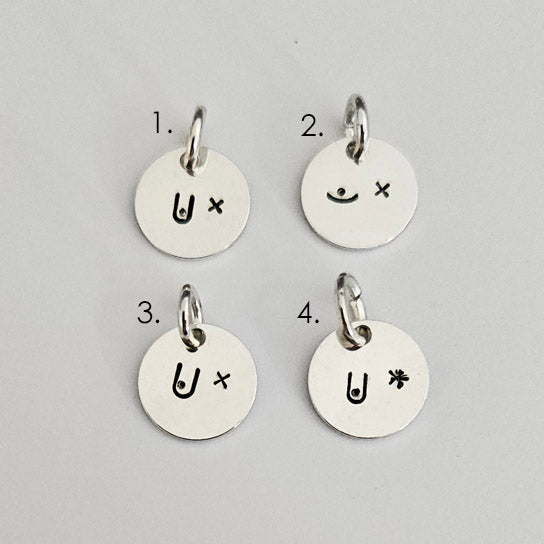 Bild på fyra runda, handstämplade berlocker i silver liggandes på en ljus bakgrund. Varje hänge har ett unikt motiv som hyllar bröstcanceröverlevare, där ena sidan visar ett bröst och andra sidan ett ärr i form av ett litet kryss eller en stjärna. Hängena är numrerade 1 till 4.