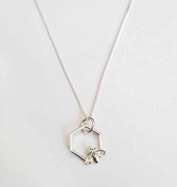 Närbild på ett silverhalsband med ett hängsmycke format som en sexkantig hexagon. I botten av ramen sitter ett litet, detaljerat bi i silver med en klar sten på ryggen.